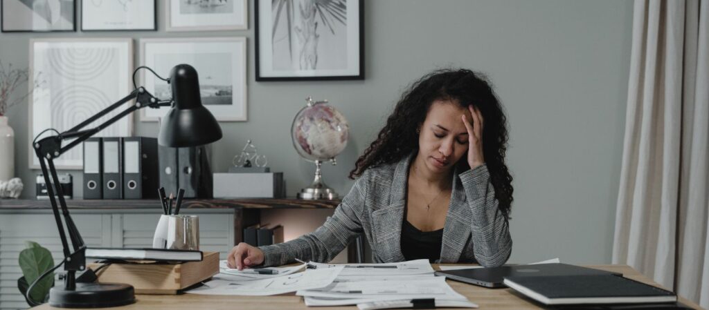 Une professeure universitaire visiblement fatiguée à son bureau, entourée de documents et d’un ordinateur. Cette image illustre les défis liés à la surcharge de travail et à l’isolement, des facteurs menaçant la collégialité et la gouvernance participative dans le milieu académique.