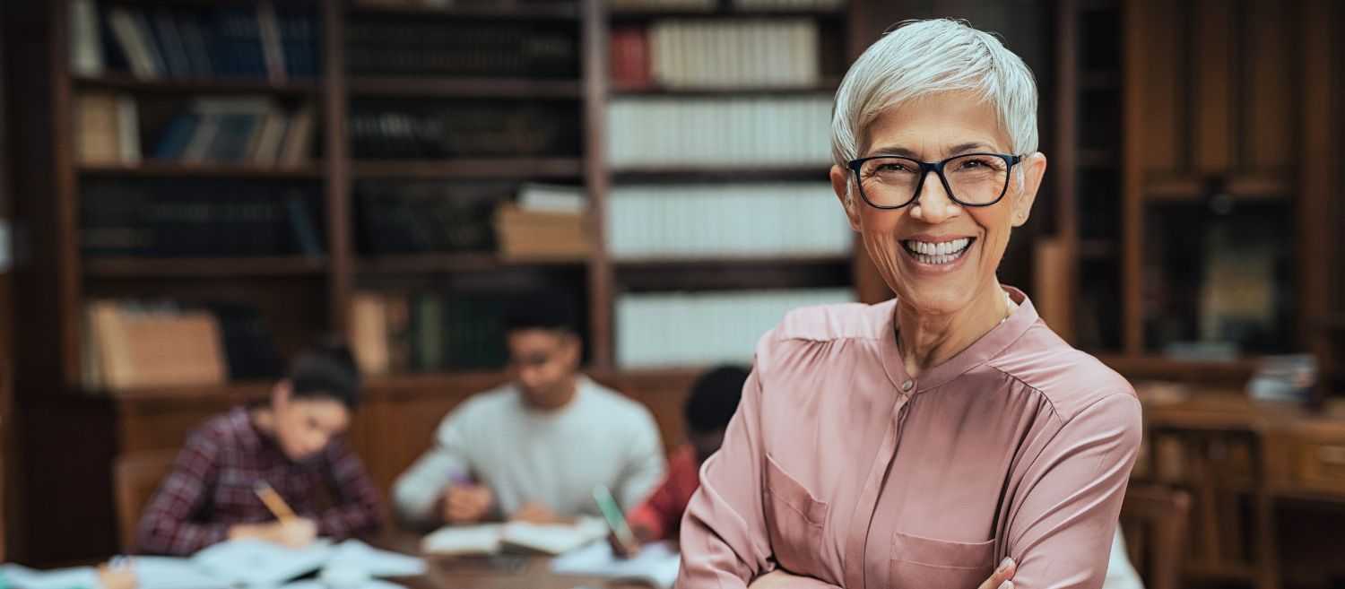 Professeure senior souriante devant une bibliothèque universitaire, représentant les avancées des luttes féministes pour l’égalité des femmes en milieu académique. Image illustrant l’engagement pour une plus grande représentation des femmes dans l’enseignement supérieur et la recherche.