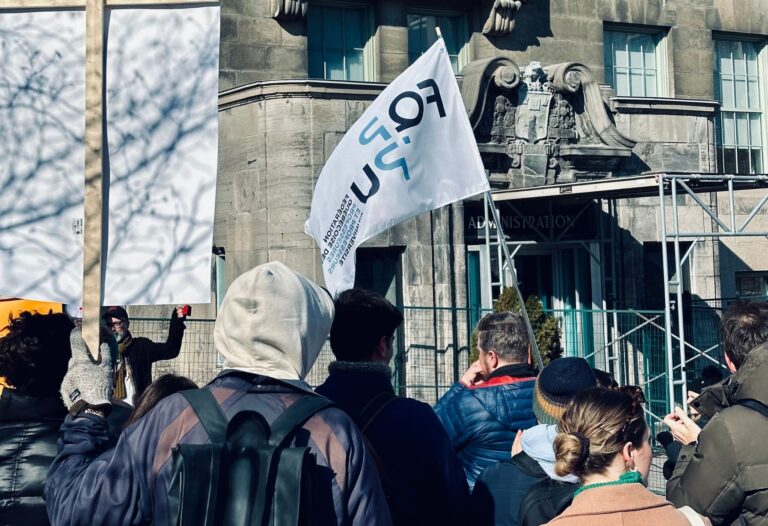 Manifestation devant un bâtiment à Montréal : des manifestants brandissent des pancartes et un drapeau de la FQPPU pour défendre la démocratie syndicale et s’opposer à la réforme du régime syndical visant les cotisations syndicales.