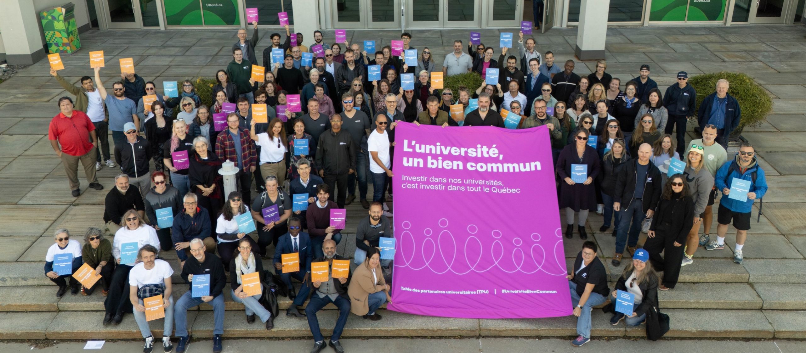 Grand groupe de membres de la communauté universitaire rassemblés autour d’une bannière « L’université, un bien commun ».