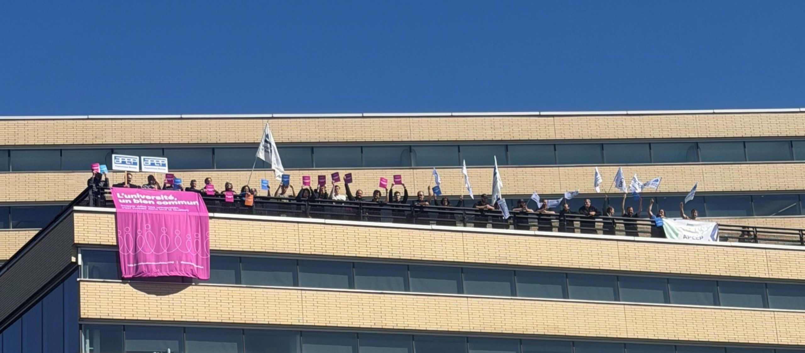 Des membres de la communauté universitaire mobilisés sur une terrasse avec une grande bannière « L’université, un bien commun ».